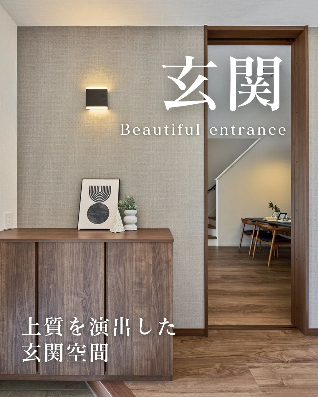 温かいミディアムブラウンの木目調シューズボックスとフローリングの玄関。壁はベージュ系の布地のような質感のアクセントクロスで、シンプルな上下配光のウォールライトと、モダンな抽象画が飾られています。