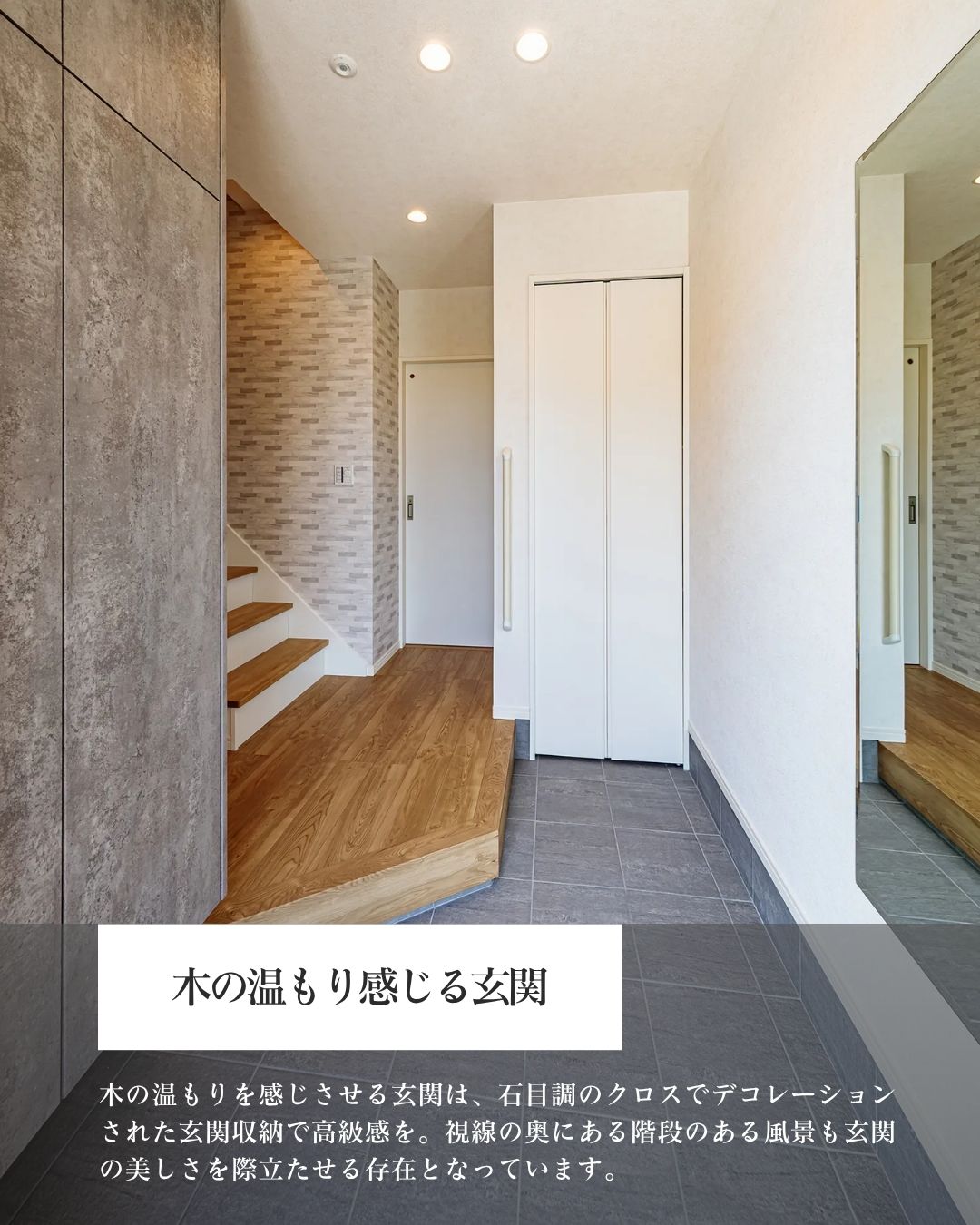 注文住宅の玄関。木目調の上がり框とフローリングが暖かさを演出しており、壁面全体にはグレー系の石目調のクロスが貼られています。右側には大容量の白い扉の玄関収納があり、全身鏡も設置されています。奥には木製の踏板と白い蹴込みの階段が見える、機能的なデザインの玄関です。