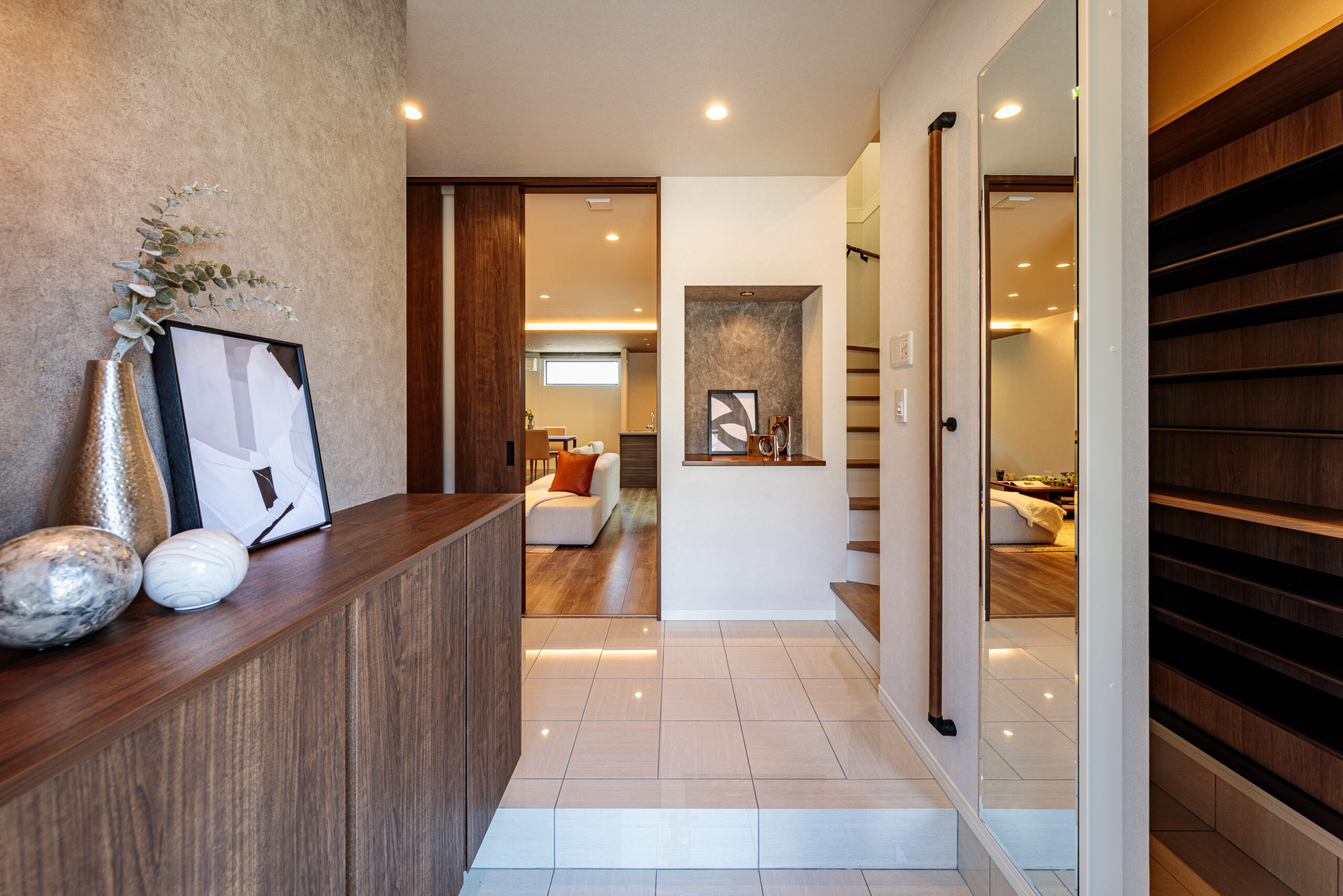 目調のフロート型シューズボックスが設置された、明るい注文住宅の玄関ホール。右手の壁には全身鏡とオープンの棚状の収納スペースがあり、実用性に優れています。正面奥の引き戸の向こうにはリビング・ダイニングが見え、正面にはニッチがあり、アートや小物が飾られています。